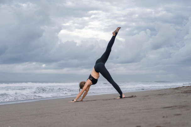 sporty young woman practicing yoga in One Legged Downward-Facing Dog pose (Eka Pada Adho Mukha Svanasana) on seashore - Foto, Imagen
