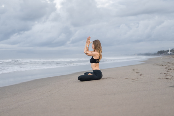 beautiful young woman practicing yoga in lotus pose on seashore on cloudy day - Photo, Image