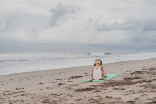 criança pequena feliz que pratica ioga no cão Upward-Facing (Urdhva Mukha Svanasana) posar na praia
 - Foto, Imagem