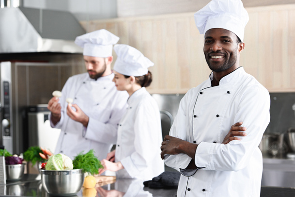 Chef afroamericano de pie con los brazos cruzados frente a sus colegas en la cocina
 - Foto, imagen