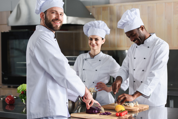 Equipo multirracial de cocineros sonriendo mientras cortan ingredientes por la mesa de la cocina
 - Foto, imagen