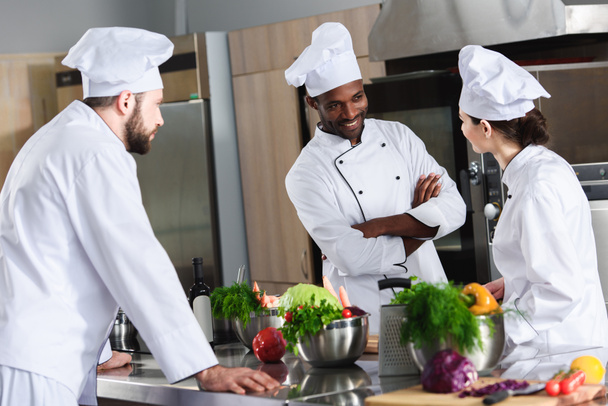 Equipo de chefs multirraciales discutiendo nueva receta por el mostrador de cocina
 - Foto, Imagen