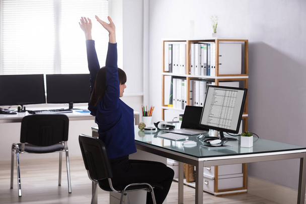 Young Businesswoman Sitting On Chair Raising Her Arms At Workplace - Фото, изображение