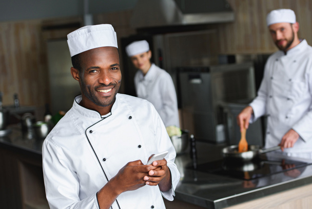 smiling african american chef using smartphone at restaurant kitchen and looking at camera - Photo, Image