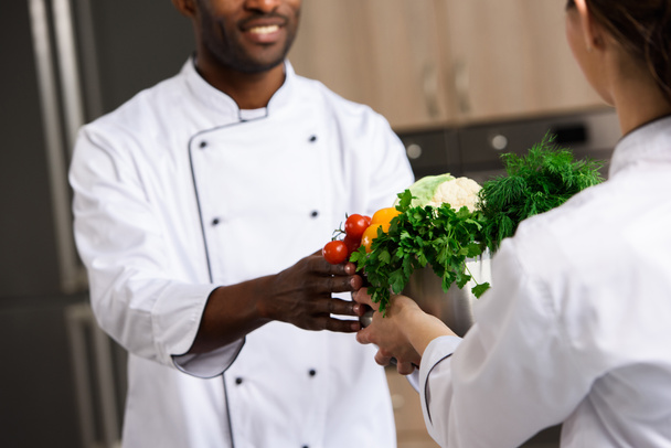 imagen recortada de chef dando tazón con verduras a un colega afroamericano en la cocina del restaurante
 - Foto, Imagen