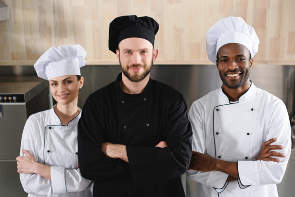 chefs multiculturales de pie con los brazos cruzados y mirando a la cámara en la cocina del restaurante
 - Foto, Imagen