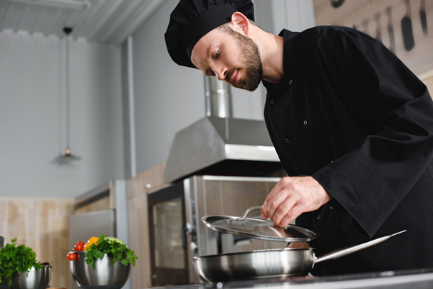 vista lateral de la cocina guapo chef con sartén en la cocina del restaurante
 - Foto, Imagen