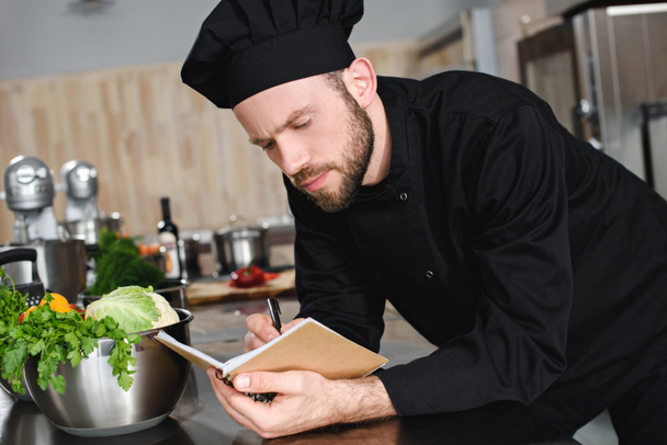 vista lateral del guapo chef escribiendo una nueva receta para portátil en la cocina del restaurante
 - Foto, Imagen