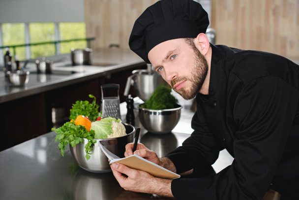 handsome chef writing down new recipe to notebook at restaurant kitchen and looking away - Photo, Image