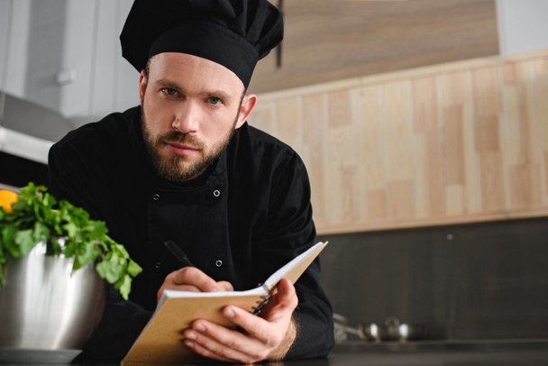guapo chef escribiendo una nueva receta para portátil en la cocina del restaurante y mirando a la cámara
 - Foto, Imagen