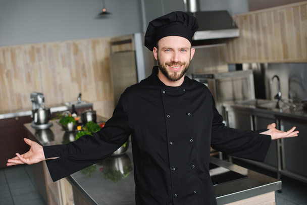sonriente guapo chef de pie con los brazos abiertos en la cocina del restaurante
 - Foto, Imagen