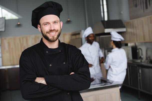 chef sonriente de pie con los brazos cruzados y mirando a la cámara en la cocina del restaurante
 - Foto, Imagen