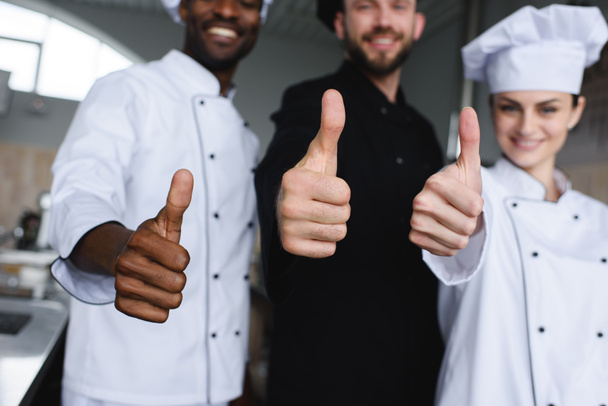 przycięty obraz uśmiechający się wielokulturowym kucharzy wyświetlane kciuki w kuchni restauracji - Zdjęcie, obraz