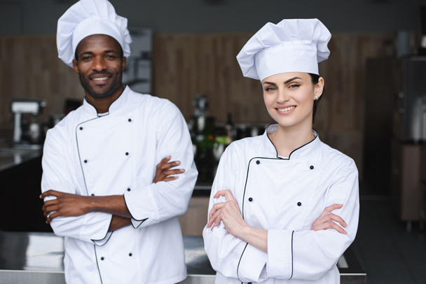 chefs multiculturales de pie con los brazos cruzados y mirando a la cámara en la cocina del restaurante
 - Foto, Imagen