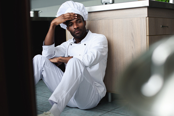 exhausto chef afroamericano sentado en el piso en la cocina del restaurante y mirando hacia otro lado
 - Foto, Imagen