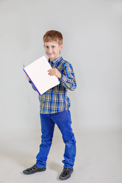 Portrait of cute Caucasian boy, elementary school student with book  on grey background. child aged 7 years first grader full-length view - Фото, изображение