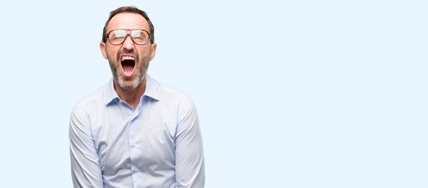 Middle age man with glasses stressful, terrified in panic, shouting exasperated and frustrated. Unpleasant gesture. Annoying work drives me crazy isolated over blue background - Photo, Image