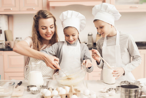 Äiti opettaa lapsilleen valkoisissa hatuissa valmistelee evästeitä keittiössä
 - Valokuva, kuva