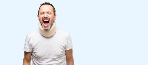Injured senior man using a neck brace stressful, terrified in panic, shouting exasperated and frustrated. Unpleasant gesture. Annoying work drives me crazy isolated over blue background - Photo, Image