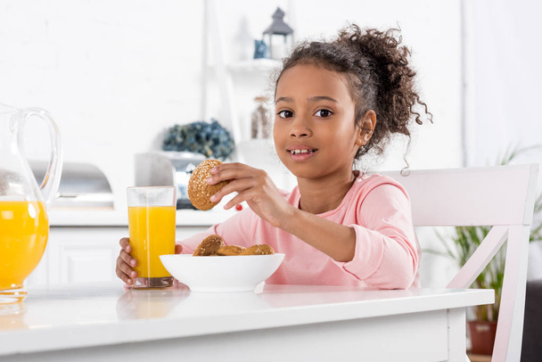 preteen african american kid with orange juice and oatmeal cookies in kitchen - Photo, Image