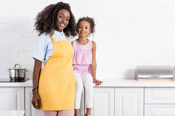 afroamericana madre en delantal e hija posando en cocina
 - Foto, imagen
