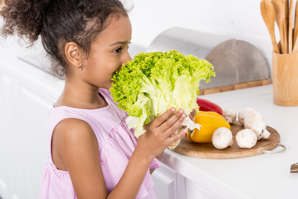 african american child sniffing green lettuce in kitchen - Foto, afbeelding