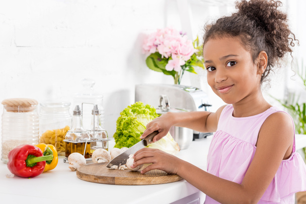 african american kid cutting vegetables on wooden board and looking at camera - 写真・画像