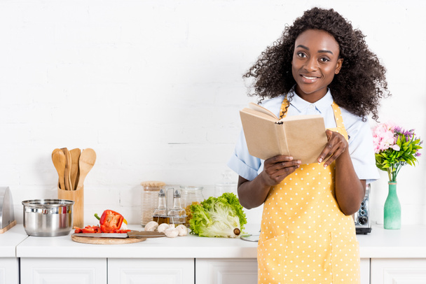 smiling african american woman in apron reading cookbook on kitchen - Valokuva, kuva
