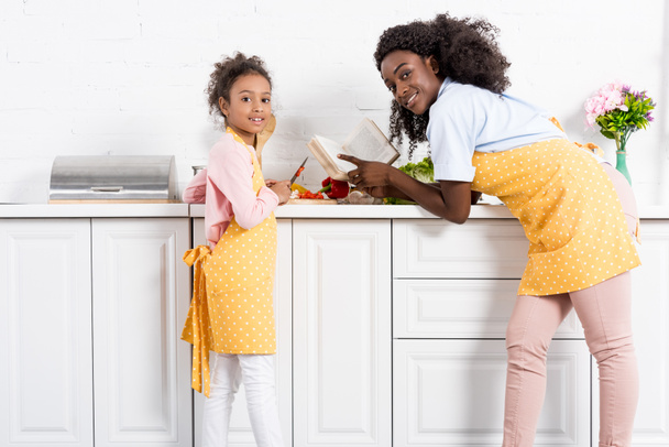 african american mother and daughter cooking with cookbook on kitchen - Zdjęcie, obraz