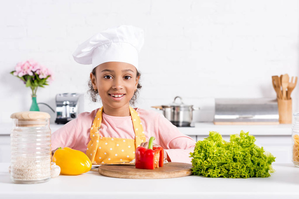 smiling african american kid in apron and chef hat preparing vegetables on kitchen  - Fotó, kép