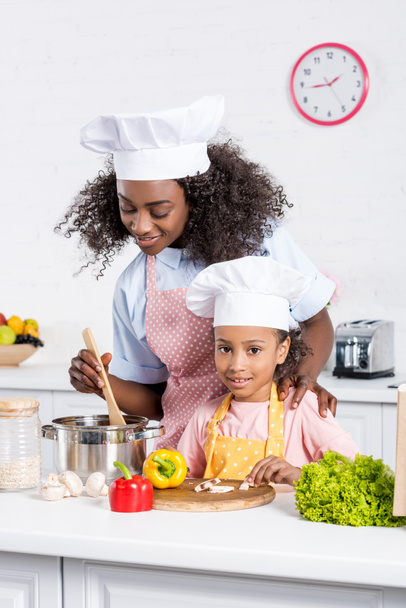 african american mother and daughter in chef hats cooking together on kitchen - Zdjęcie, obraz