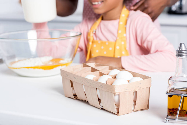 cropped view of african american kid in apron cooking on kitchen, eggs and oil standing on table on foreground - 写真・画像