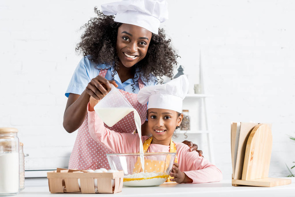 african american mother and daughter in chef hats pouring milk for dough on kitchen - Fotoğraf, Görsel