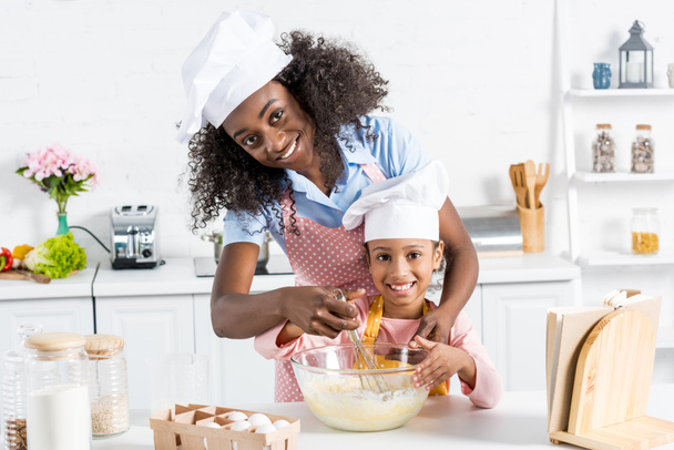 africano americano madre e hija en chef sombreros mezcla de masa con batidor en cocina
 - Foto, Imagen