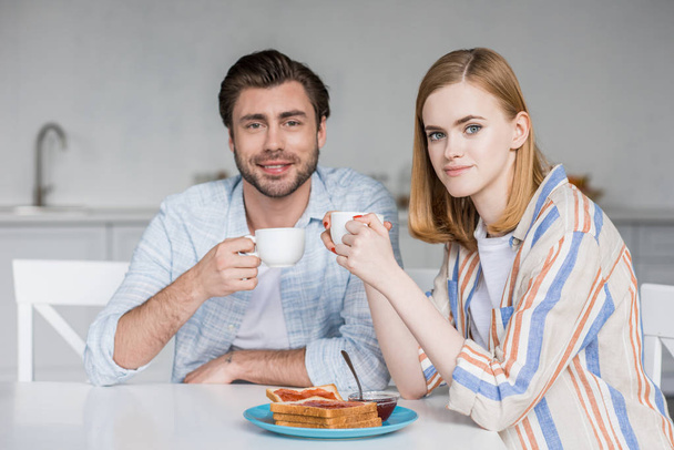 smiling young couple with coffee cups having breakfast at table in kitchen - Фото, изображение
