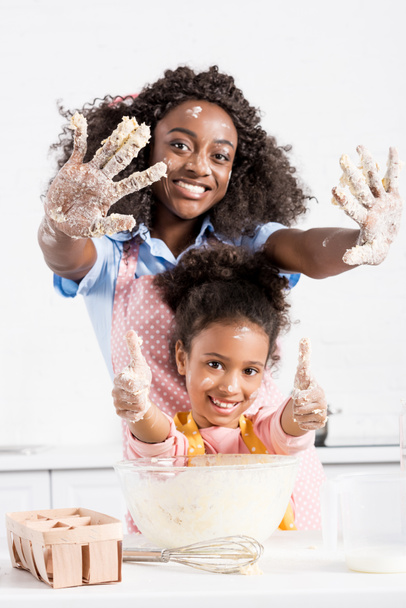 smiling african american mother and funny daughter showing hands in dough and thumbs up on kitchen - Φωτογραφία, εικόνα