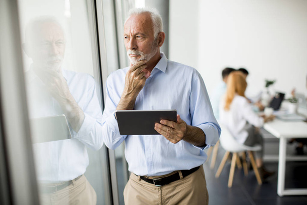 Портрет старшего бизнесмена, стоящего у окна с цифровой табличкой в руке
 - Фото, изображение