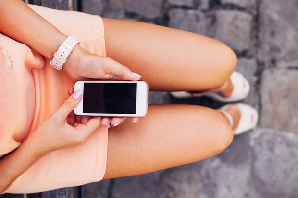 beautiful legs of a girl who sitting on the bench and holding the phone in her hands - Foto, afbeelding