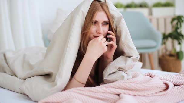 Cheerful young red haired girl lying under blanket and talk on mobile phone in bed - Filmati, video