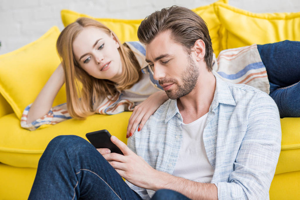 young woman laying on couch and watching boyfriend using smartphone  - Фото, изображение