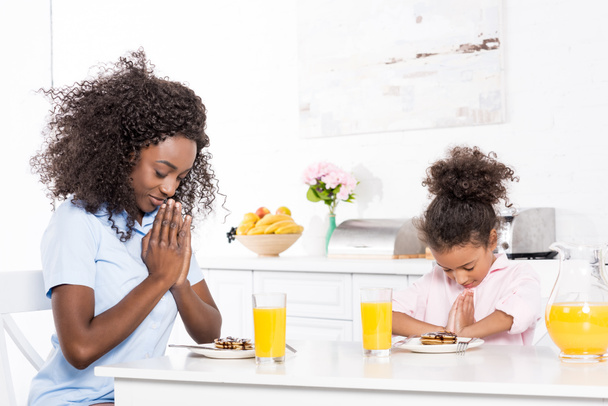 Afro-Amerikaanse moeder en dochter bidden voor het ontbijt in de keuken - Foto, afbeelding
