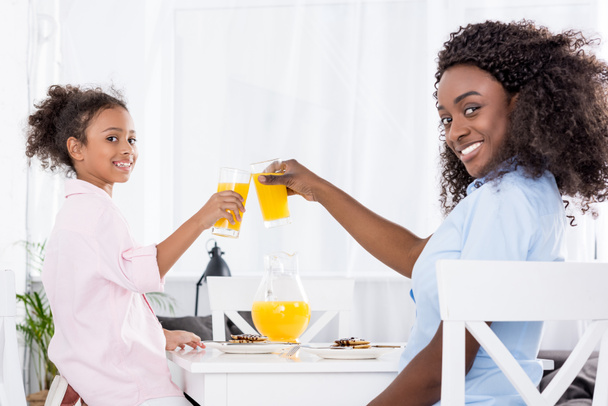 χαμογελώντας αφρικανική αμερικανική μητέρα και κόρη τσούγκρισμα με χυμό πορτοκαλιού  - Φωτογραφία, εικόνα