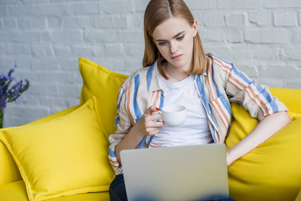 fiatal nő, kávéscsésze használ laptop, és ül a kanapén - Fotó, kép