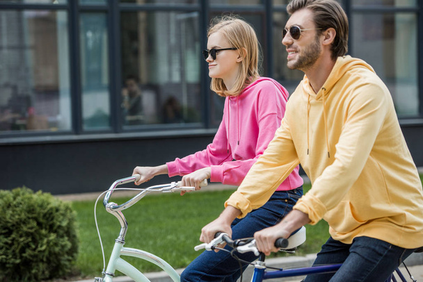 side view of stylish couple in sunglasses riding on bicycles  - Foto, imagen