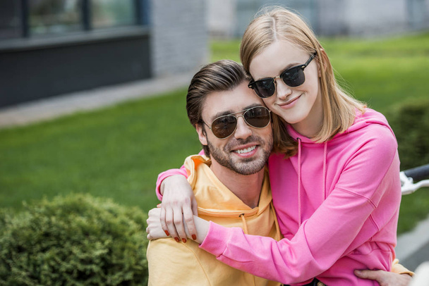 smiling young woman in sunglasses sitting on boyfriend knees and embracing him  - Photo, Image