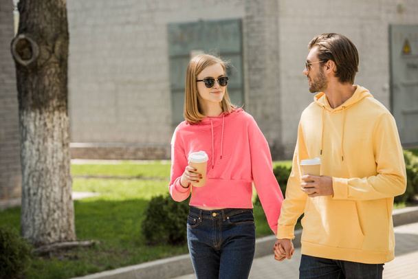 stylish young couple in sunglasses with coffee cups holding hands and walking on street - Фото, зображення