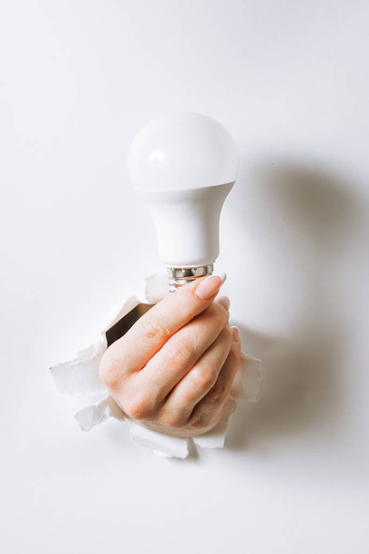 Hand holding a light bulb breaking through the hole in white bac - Photo, Image