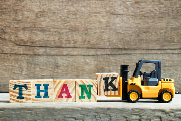 Yellow toy forklift hold letter block K to complete word thank on wood background - Foto, Imagem