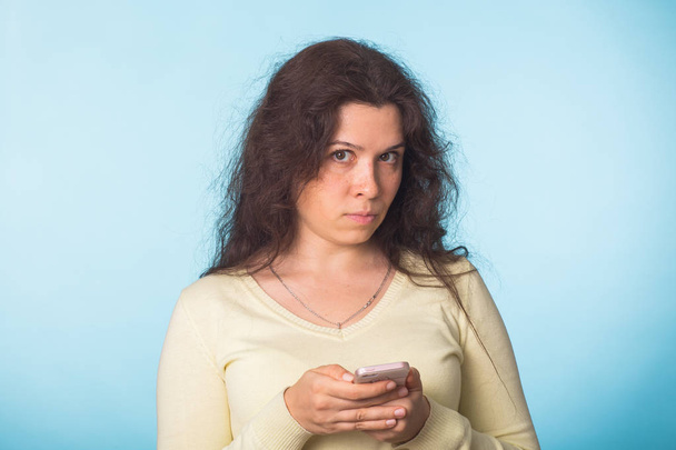 Mujer infeliz resentida sosteniendo el teléfono inteligente mirando la cámara sobre fondo azul
 - Foto, imagen