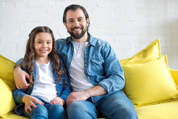 幸せな父と娘黄色のソファーに一緒に座って、カメラで笑顔  - 写真・画像